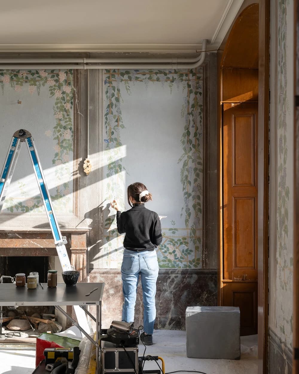 Une conservatrice-restauratrice traite une peinture murale en trompe-l’œil au Domaine de La Doges, dans un intérieur historique éclairé par le soleil.